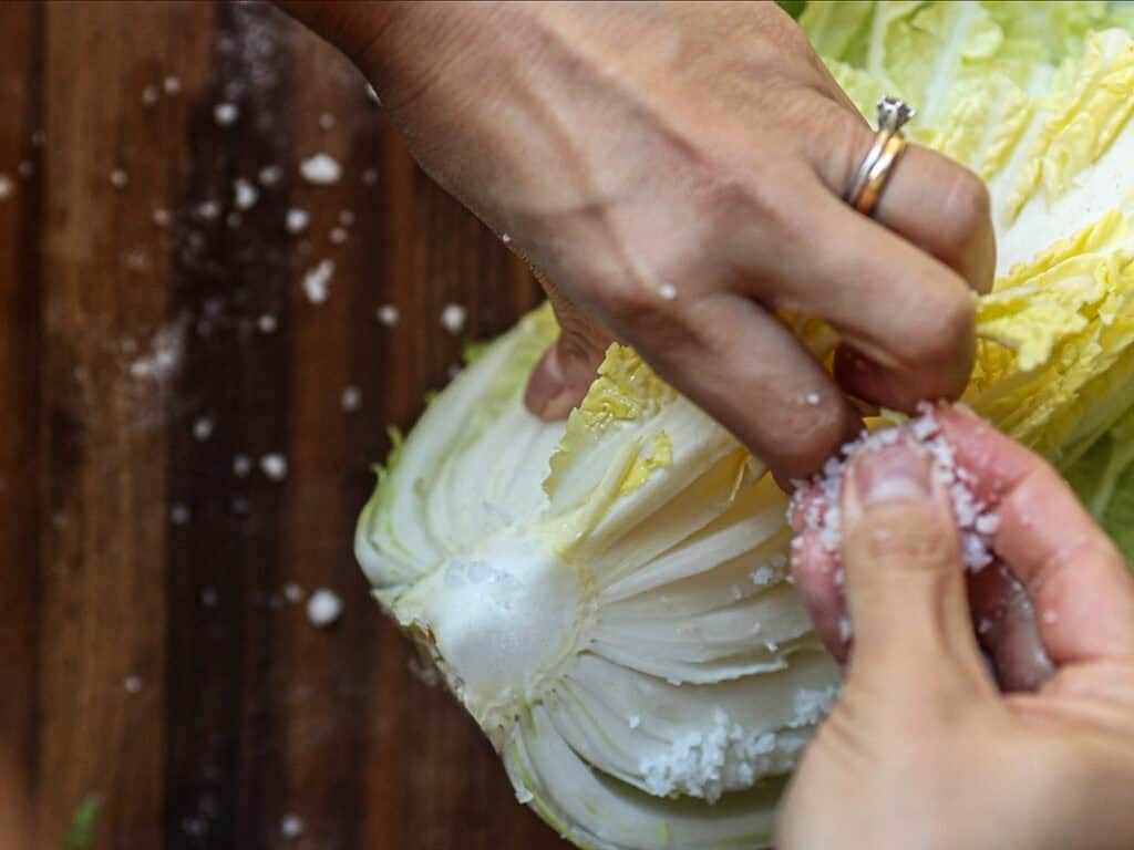 Hands are salting a half-cut napa cabbage over a wooden surface. A persons fingers are sprinkling coarse salt onto the layers of the vegetable.